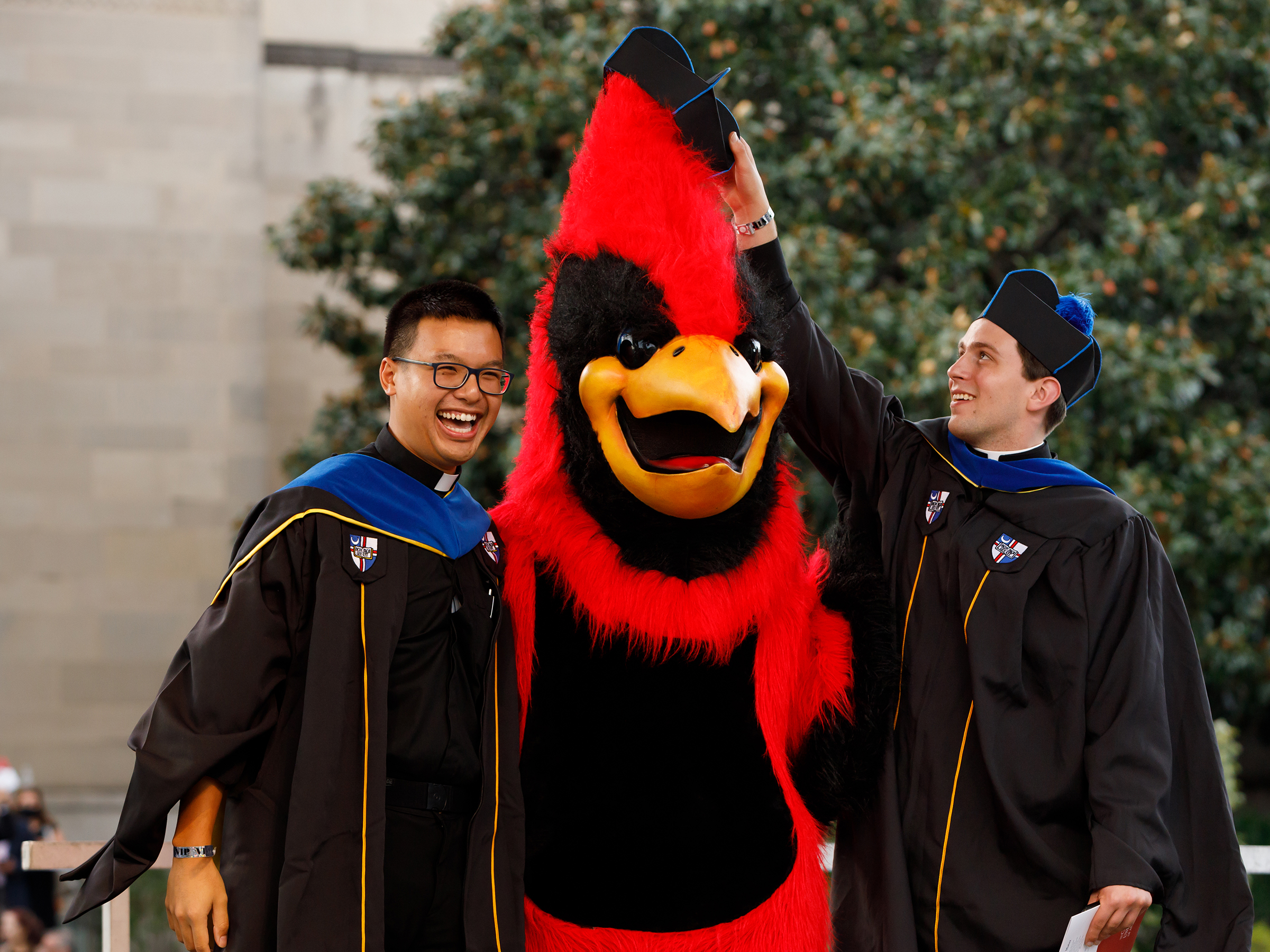 Graduates in regalia smile and put a graduate hat on Red, the mascot